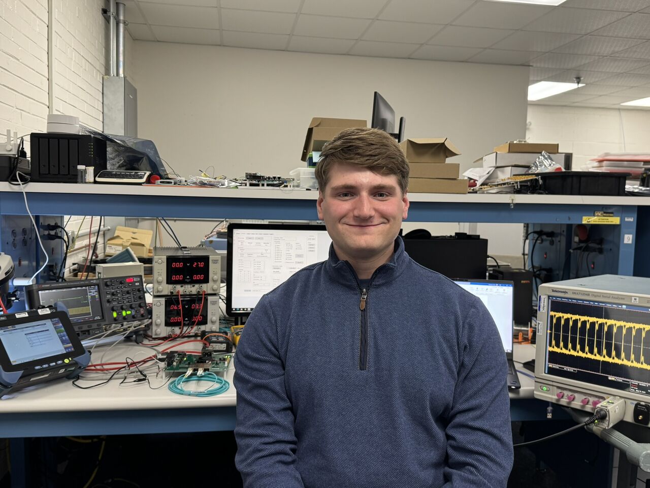 Aaron Grossman in an electronics lab beside Ethernet test equipment and an oscilloscope